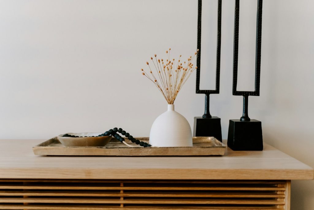 A white ceramic vase with dried flowers beside two black candle holders on a wooden tray