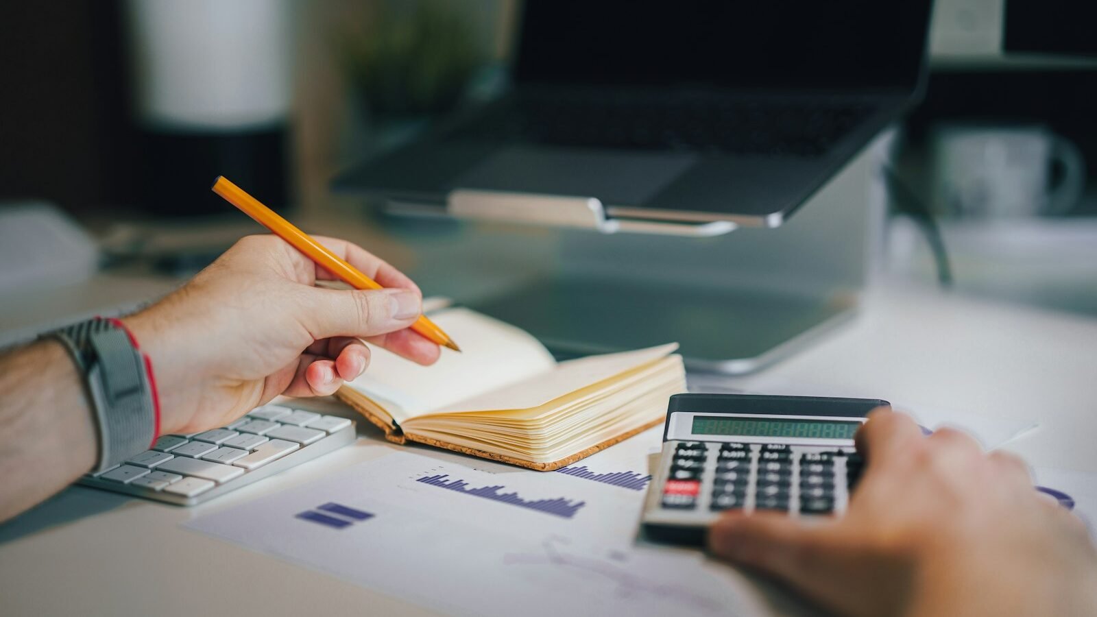 Hands holding a pencil over a notebook and pressing a calculator, with printed charts on the desk.