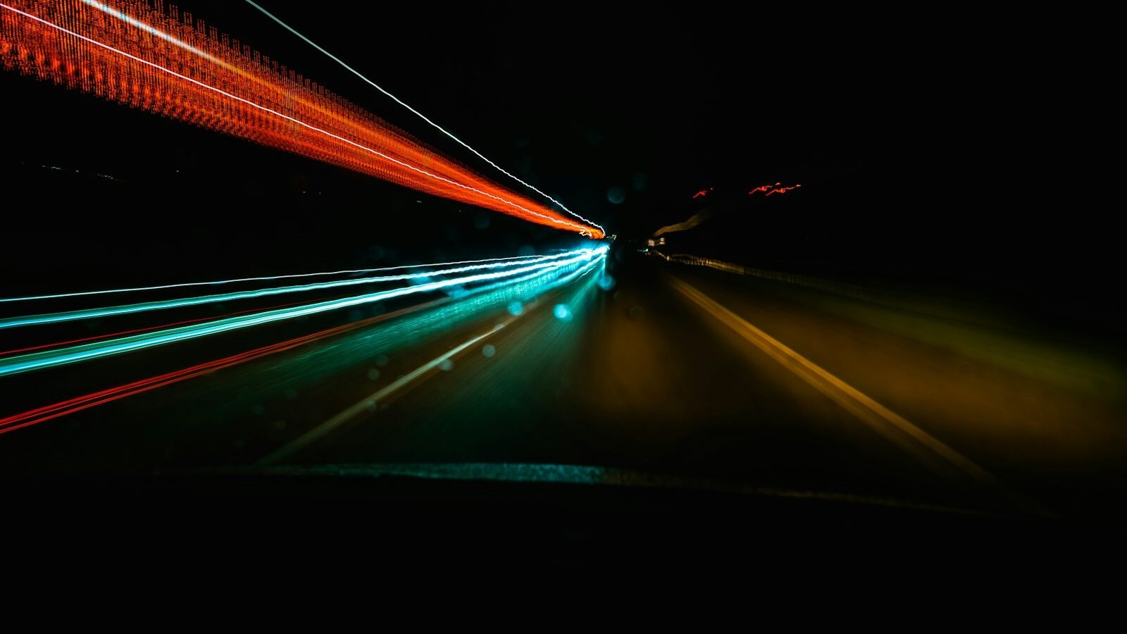 Long-exposure light trails in red and teal streaking along a night road, suggesting speed.