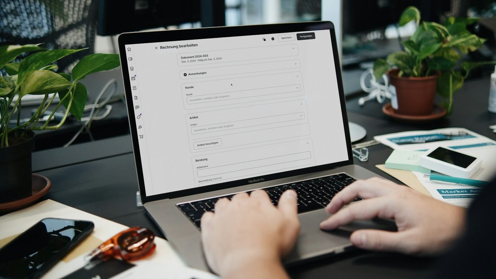 A person using a laptop to edit a form on screen, at a busy desk with plants and paperwork.