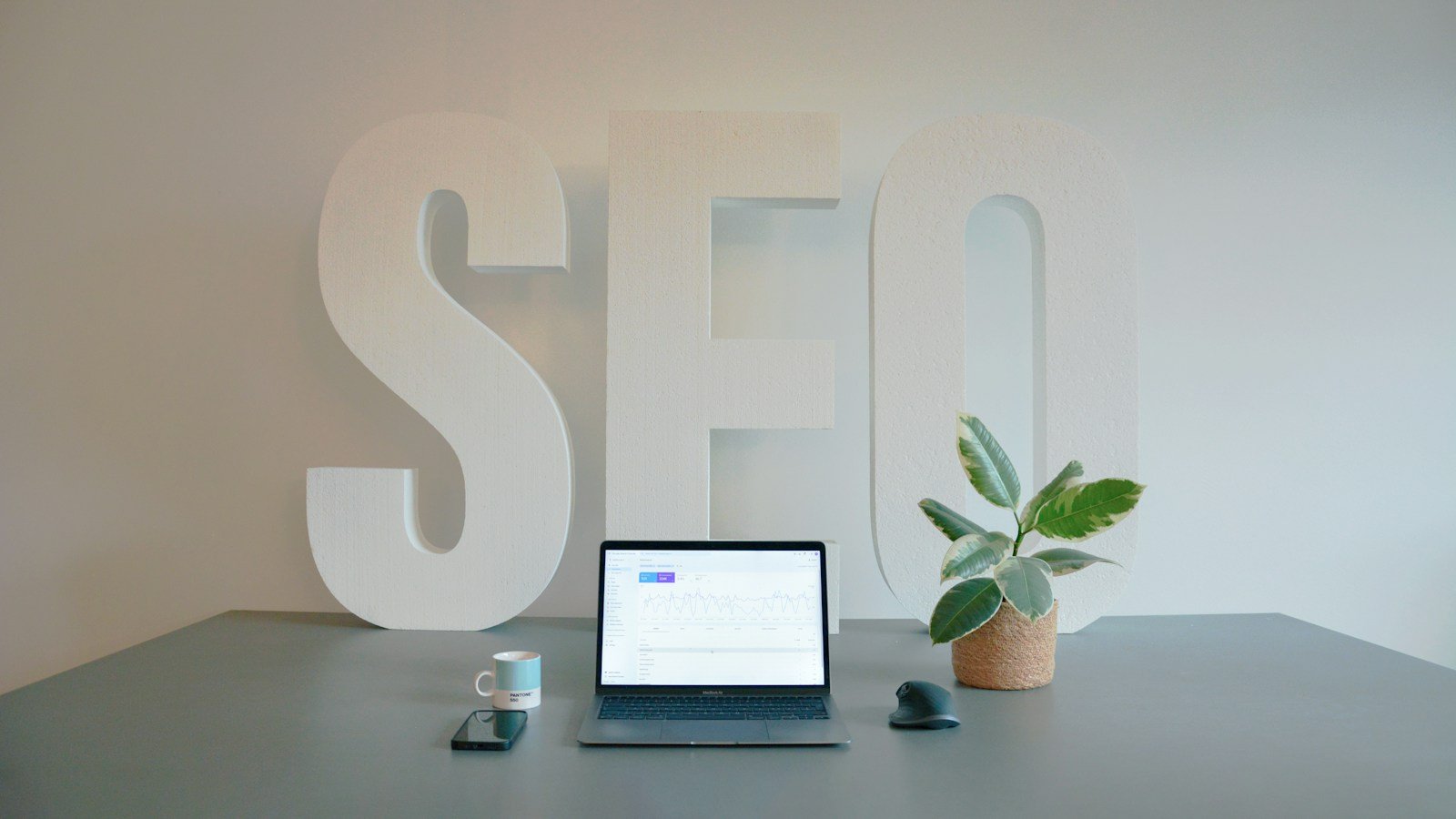 Large white letters spelling SEO on a wall behind a desk with a laptop showing a search performance chart, a coffee cup, a phone and a plant.